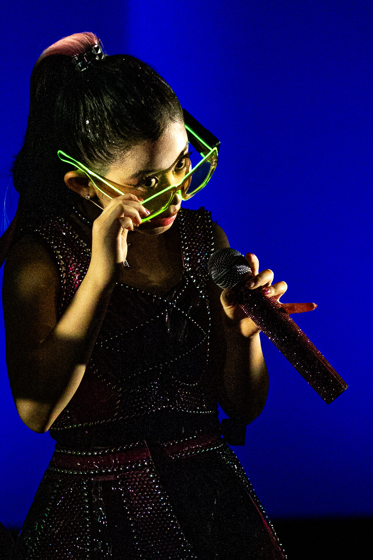 Performer in sequinned costume and light-up glasses singing on stage, Six: Teen Edition